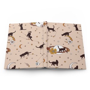 May include: An open notebook with a pink background and a repeating pattern of black cats, stars, moons, and planets. The cats are in various poses, including sitting, standing, and jumping. The stars are gold and the moons are white with a gold crescent. The planets are brown with gold rings.