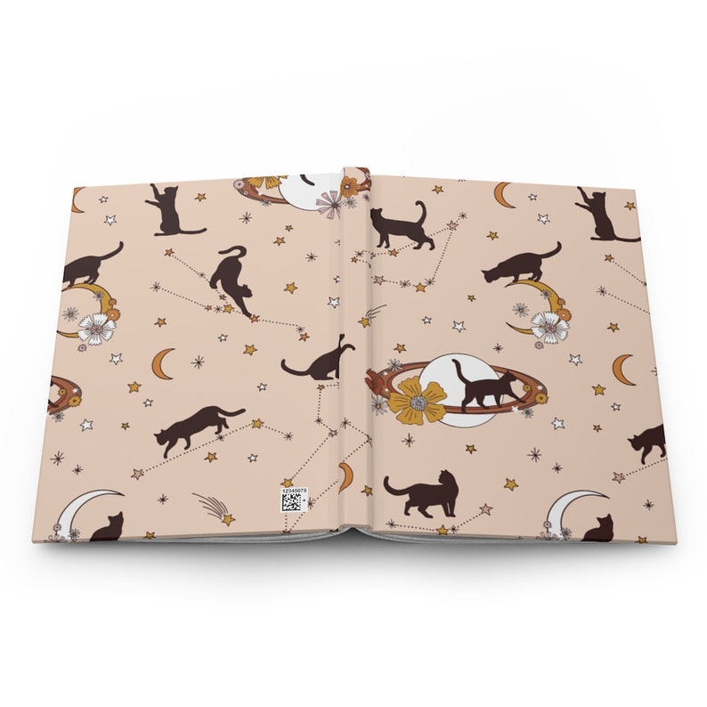 May include: An open notebook with a pink background and a repeating pattern of black cats, stars, moons, and planets. The cats are in various poses, including sitting, standing, and jumping. The stars are gold and the moons are white with a gold crescent. The planets are brown with gold rings.