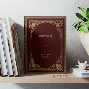 May include: A burgundy guest book with gold ornate detailing and the text "GUEST BOOK", "Jen & Quinn", and "03.12.2024". The book is on a wooden shelf with other books and a hanging plant.