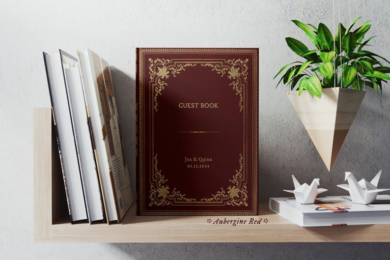 May include: A burgundy guest book with gold ornate detailing and the text "GUEST BOOK", "Jen & Quinn", and "03.12.2024". The book is on a wooden shelf with other books and a hanging plant.
