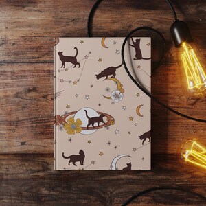 May include: A beige notebook with a celestial pattern featuring black cats, stars, moons, and planets. The notebook is on a wooden surface with two Edison bulb lights hanging from black cords.