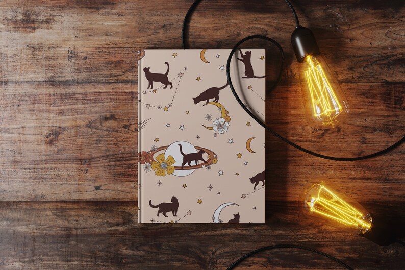 May include: A beige notebook with a celestial pattern featuring black cats, stars, moons, and planets. The notebook is on a wooden surface with two Edison bulb lights hanging from black cords.