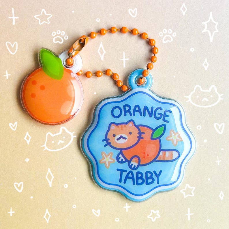 May include: A set of two orange-themed charms. One charm is an orange fruit with a green leaf. The other is a blue charm with an orange tabby cat illustration and the words "ORANGE TABBY". Both charms are connected by an orange beaded chain.