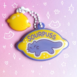 May include: A keychain featuring a yellow lemon charm and a larger lemon-shaped charm with the text "SOURPUSS" and a purple cat illustration. The keychain has a white beaded chain and a star-shaped clasp.