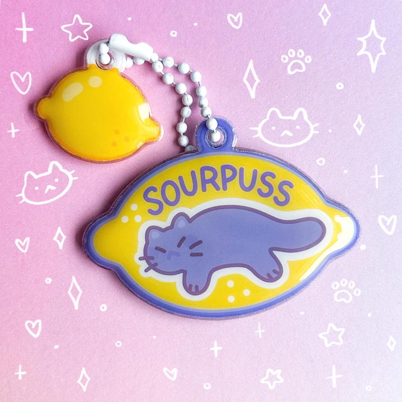 May include: A keychain featuring a yellow lemon charm and a larger lemon-shaped charm with the text "SOURPUSS" and a purple cat illustration. The keychain has a white beaded chain and a star-shaped clasp.