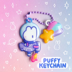 Puede incluir: Un llavero de astronauta morado y azul con una estrella fugaz arcoíris y el texto "PUFFY KEYCHAIN".