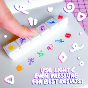 May include: A white stamp set with various colorful icons, including a crown, potion, sword, and shield. The text "USE LIGHT & EVEN PRESSURE FOR BEST RESULTS!" is also visible. A finger presses down on one of the stamps.