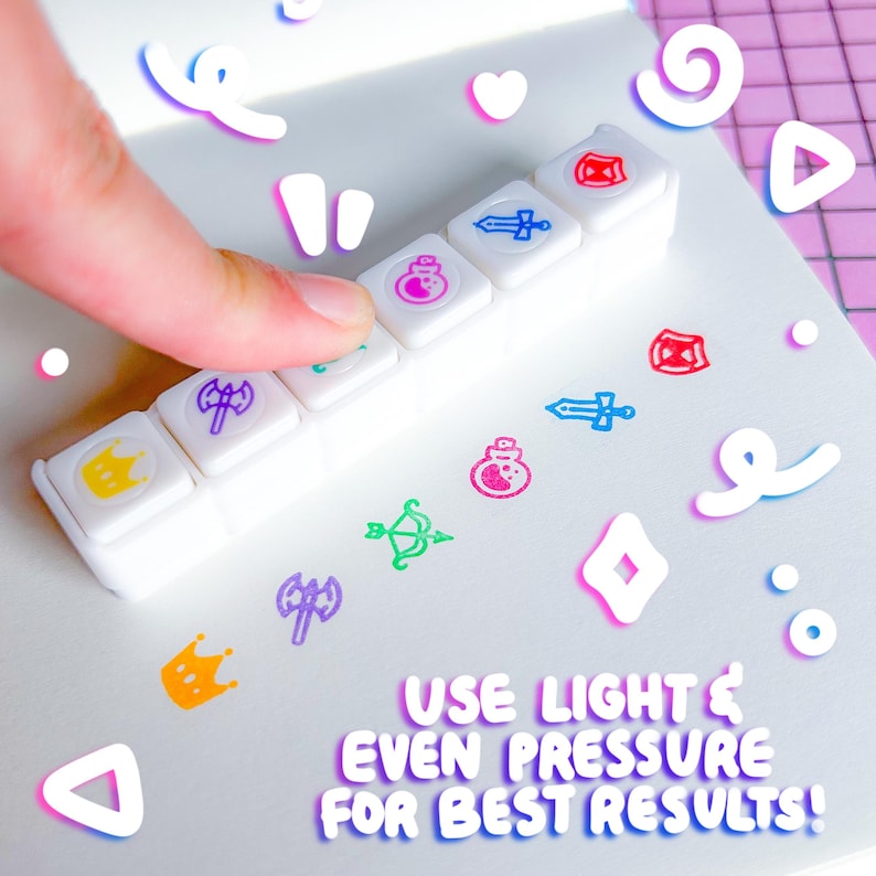 May include: A white stamp set with various colorful icons, including a crown, potion, sword, and shield. The text "USE LIGHT & EVEN PRESSURE FOR BEST RESULTS!" is also visible. A finger presses down on one of the stamps.