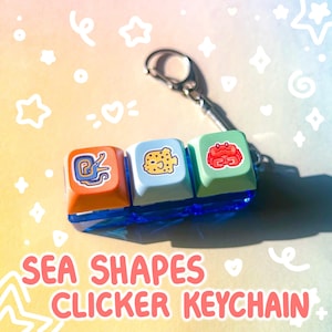 May include: A colorful keychain featuring three clicker keycaps with sea creature designs: a snail, a spotted fish, and a crab. The keycaps are orange, light blue, and green, attached to a silver keyring. The text "SEA SHAPES CLICKER KEYCHAIN" is at the bottom.