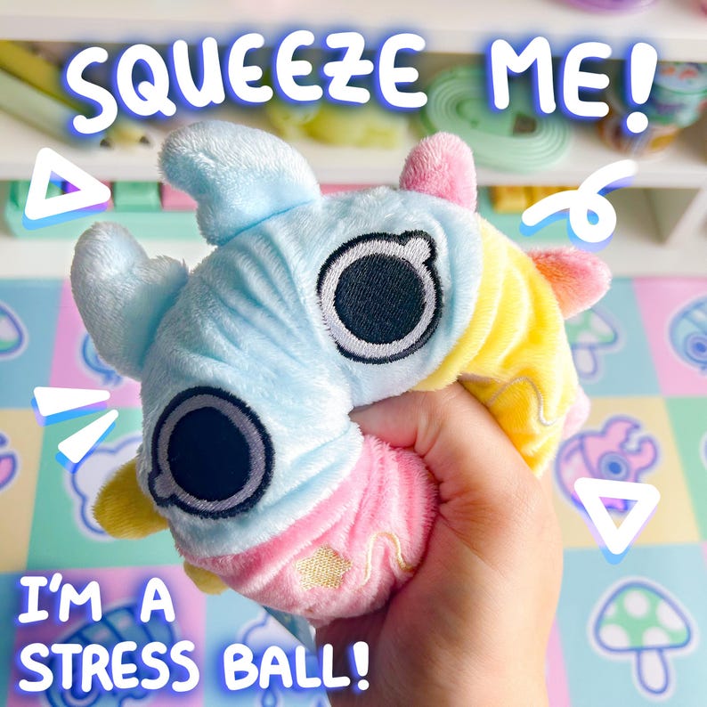 Puede incluir: Un juguete de peluche colorido con forma de gusano con ojos grandes. El juguete est&aacute; siendo apretado en una mano. El texto "Squeeze Me!" est&aacute; escrito encima del juguete y "I'm a stress ball!" est&aacute; escrito debajo del juguete.