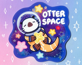 May include: A die-cut sticker featuring a cartoon otter wearing a space helmet and an orange suit, floating in a dark blue glittery galaxy background. The text OTTER SPACE is printed in white letters. Several colorful stars surround the otter.