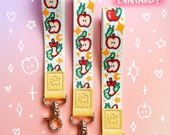 Op de afbeelding: Drie gele en witte polsbandjes met een herhalend patroon van rode appels, groene wormen en gele sterren. Elk polsbandje heeft een gele leren band met een stervormig ontwerp. De polsbandjes bevinden zich op een roze achtergrond met witte harten, sterren en plustekens. "WRIST LENGTH LANYARD!" staat in witte tekst op een roze achtergrond.