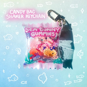 May include: A clear candy bag shaker keychain with the text "CANDY BAG SHAKER KEYCHAIN" and "SEA BUNNY GUMMIES". The bag contains colourful gummy candies and glitter, and has a gold-coloured clasp.