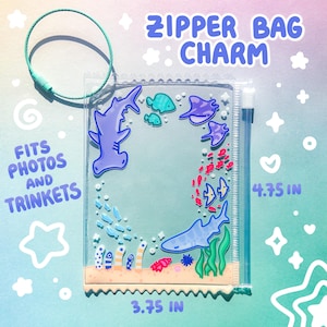May include: Clear zipper bag charm with a marine life design. The charm features a hammerhead shark, stingrays, and other sea creatures. The bag is 3.75 inches wide and 4.75 inches tall. The text "ZIPPER BAG CHARM" and "FITS PHOTOS AND TRINKETS" are visible.