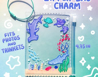 May include: Clear zipper bag charm with a marine life design. The charm features a hammerhead shark, stingrays, and other sea creatures. The bag is 3.75 inches wide and 4.75 inches tall. The text "ZIPPER BAG CHARM" and "FITS PHOTOS AND TRINKETS" are visible.