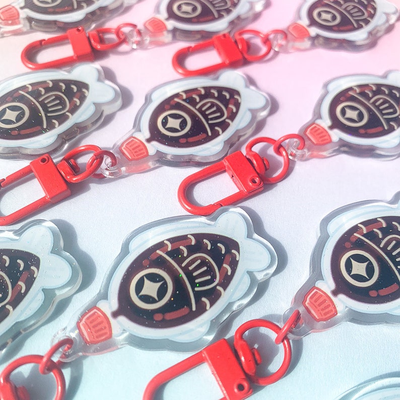 May include: Clear acrylic keychains with a fish-shaped design, featuring brown and white details, and a red clasp. The keychains have a glittery effect and are arranged on a gradient pink and blue background.