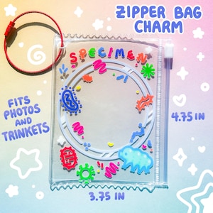 以下が含まれることがあります： カラフルな漫画風イラストと「SPECIMEN」の文字が入った、透明なプラスチック製のジッパーバッグチャーム。チャームの幅は9.5cm、高さは12.1cmです。赤いワイヤーキーチェーンが上部に取り付けられています。バッグのテキストは「FITS PHOTOS AND TRINKETS」です。