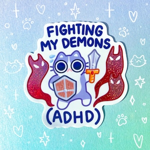 May include: A sticker featuring a cartoon cat with a shield and sword, with the text "FIGHTING MY DEMONS (ADHD)". The cat is light purple and white, with large blue eyes. Two red glittery demon figures flank the cat. The background is light blue with hearts and paw prints.