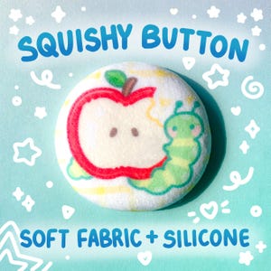 May include: A round, white squishy button with a cartoon apple design being eaten by a green caterpillar. The button has the text "SQUISHY BUTTON" and "SOFT FABRIC + SILICONE" in blue. The background is light blue with white stars and swirls.