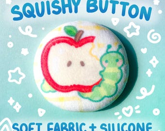 Könnte beinhalten: Runder, weißer Squishy-Button mit einem Cartoon-Apfel-Design, der von einer grünen Raupe gefressen wird. Der Button trägt den Text "SQUISHY BUTTON" und "SOFT FABRIC + SILICONE" in Blau. Der Hintergrund ist hellblau mit weißen Sternen und Wirbeln.