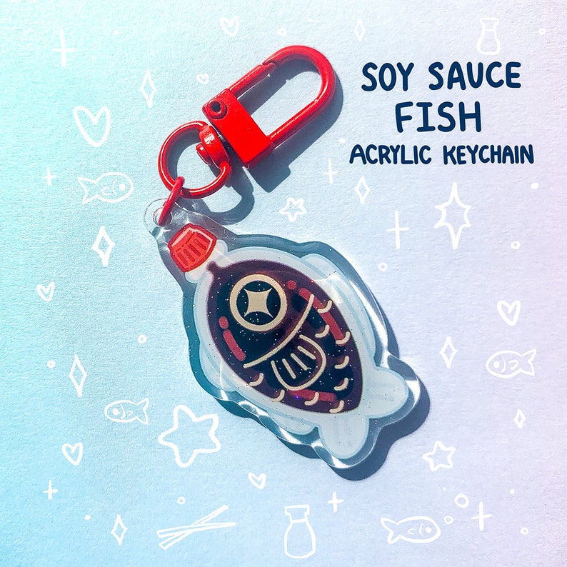 May include: An acrylic keychain shaped like a soy sauce fish, with a red clasp. The fish design features a dark brown body with red and gold accents, and the text "SOY SAUCE FISH ACRYLIC KEYCHAIN" is displayed in blue.