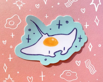 May include: A sticker of a blue and white manta ray with a yellow yolk in the center, resembling a fried egg. The sticker is on a pink background with white hearts, stars, and plus signs.