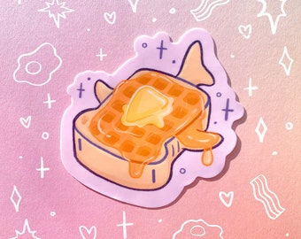 May include: A sticker of a waffle shaped like a shark with a pat of butter on top and syrup dripping down the sides. The sticker is on a pink and purple background with white hearts, stars, and crosses.