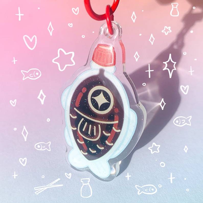 May include: A clear acrylic keychain shaped like a soy sauce bottle with a red cap. The bottle is filled with a dark brown liquid and has a white fish-shaped outline. The keychain is attached to a red ring.