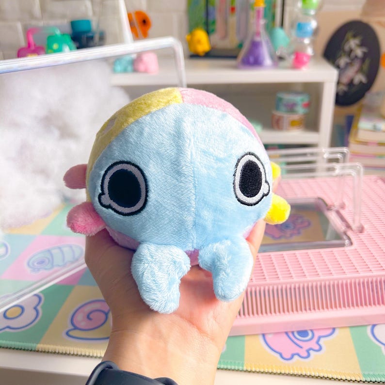 Puede incluir: Un juguete de peluche con forma de criatura redonda y colorida con grandes ojos negros y dos peque&ntilde;os brazos. El juguete est&aacute; hecho de tela suave azul, rosa y amarilla.