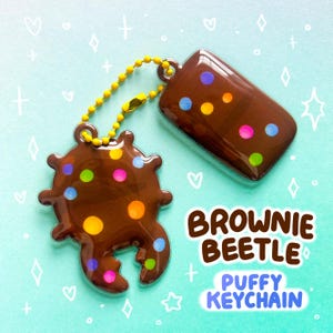 Puede incluir: Un juego de llaveros hinchados marrones con forma de escarabajo brownie, con lunares de colores. El llavero tiene una cadena dorada y una etiqueta rectangular. El texto de la imagen dice "BROWNIE BEETLE PUFFY KEYCHAIN."