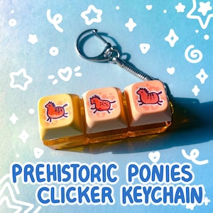 May include: A keychain featuring three clicker keycaps with a prehistoric pony design. The keycaps are yellow and peach-colored, with a red and black pony illustration. The text "PREHISTORIC PONIES CLICKER KEYCHAIN" is displayed in blue.