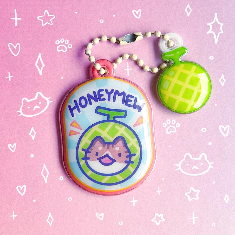 May include: A colorful keychain featuring a cat inside a melon design, with the word "HONEYMEW" above. The keychain has a pink, blue, green, and orange color scheme, with a small melon charm attached. The background is pink with white doodles.