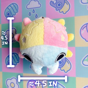 Puede incluir: Un juguete de peluche con forma de criatura redonda con un esquema de color azul claro, rosa y amarillo. El juguete tiene un dise&ntilde;o de estrella en las secciones rosa y amarilla y ojos negros. El juguete tiene aproximadamente 4,5 pulgadas de di&aacute;metro.
