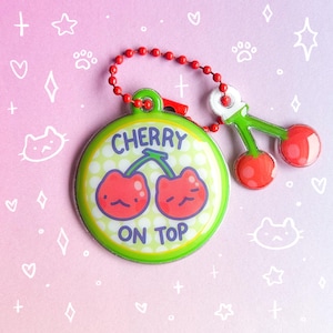 May include: A round, green keychain with the text "CHERRY ON TOP" and a cartoon cherry design. The keychain has a red beaded chain and a small cherry charm. The background is pink and purple with white doodles.