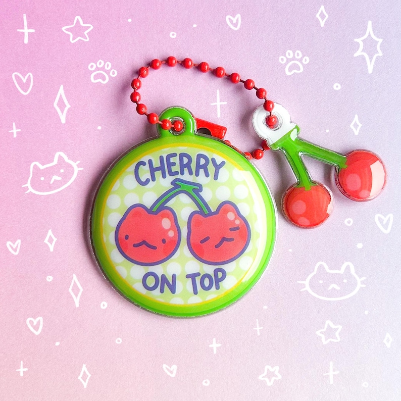 May include: A round, green keychain with the text "CHERRY ON TOP" and a cartoon cherry design. The keychain has a red beaded chain and a small cherry charm. The background is pink and purple with white doodles.