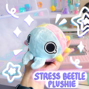 Puede incluir: Un juguete de peluche azul y rosa con forma de escarabajo, con la cara amarilla y los ojos negros. El juguete est&aacute; etiquetado como "Stress Beetle Plushie".