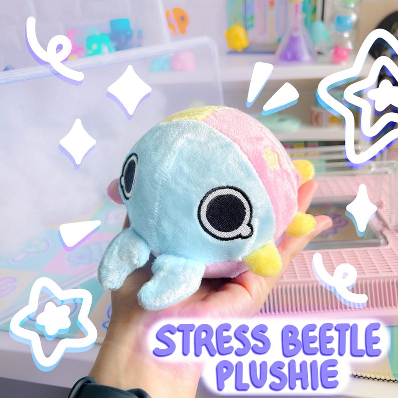 Puede incluir: Un juguete de peluche azul y rosa con forma de escarabajo, con la cara amarilla y los ojos negros. El juguete est&aacute; etiquetado como "Stress Beetle Plushie".