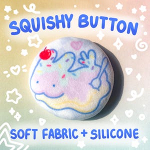 May include: A round, plush squishy button with a pastel design. The button features a light blue outline of a bunny, sprinkles, and a cherry on top. The text "SQUISHY BUTTON" and "SOFT FABRIC + SILICONE" are displayed in blue.