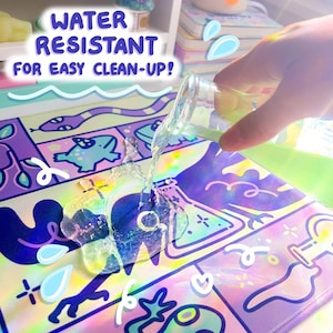 Puede incluir: Una alfombrilla de escritorio hologr&aacute;fica colorida con un dise&ntilde;o caprichoso que presenta ranas, plantas y vasos de precipitados. La alfombrilla es resistente al agua, lo que facilita la limpieza de los derrames. El texto "WATER RESISTANT FOR EASY CLEAN-UP!" est&aacute; escrito en una fuente l&uacute;dica.