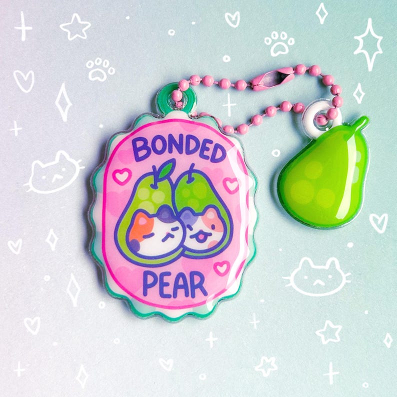 May include: A pink and green keychain with a pear and cat design. The keychain features a pink tag with the words "Bonded Pear" and two cartoon cats inside pears. A green pear charm hangs from a pink beaded chain.