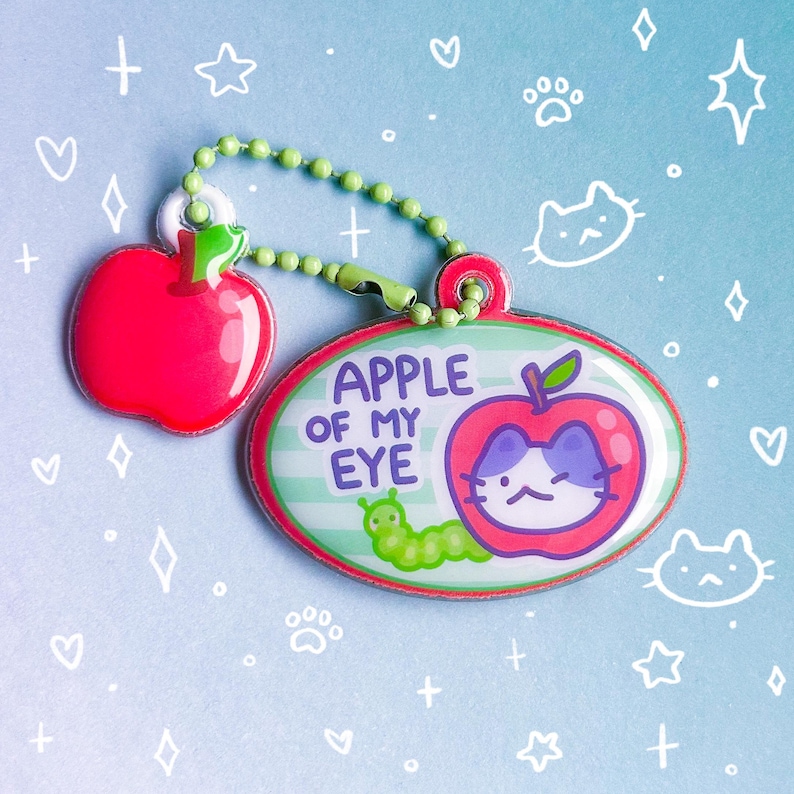 May include: A keychain with two charms: a red apple and an oval charm with the text "APPLE OF MY EYE". The oval charm features a cat inside an apple and a green caterpillar. The keychain has a green beaded chain.
