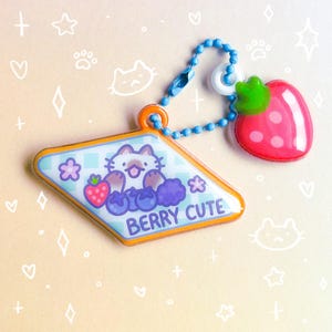 May include: A charm with a cat illustration surrounded by blueberries, strawberries, and flowers. The charm has the text "BERRY CUTE" and is connected to a strawberry charm with a blue beaded chain.