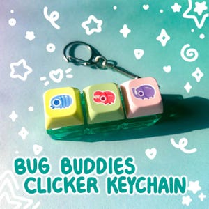 May include: A keychain featuring three colorful keycaps with bug illustrations. The keycaps are yellow, green, and pink, each with a different bug design. The keychain has a silver metal ring and the text "BUG BUDDIES CLICKER KEYCHAIN".