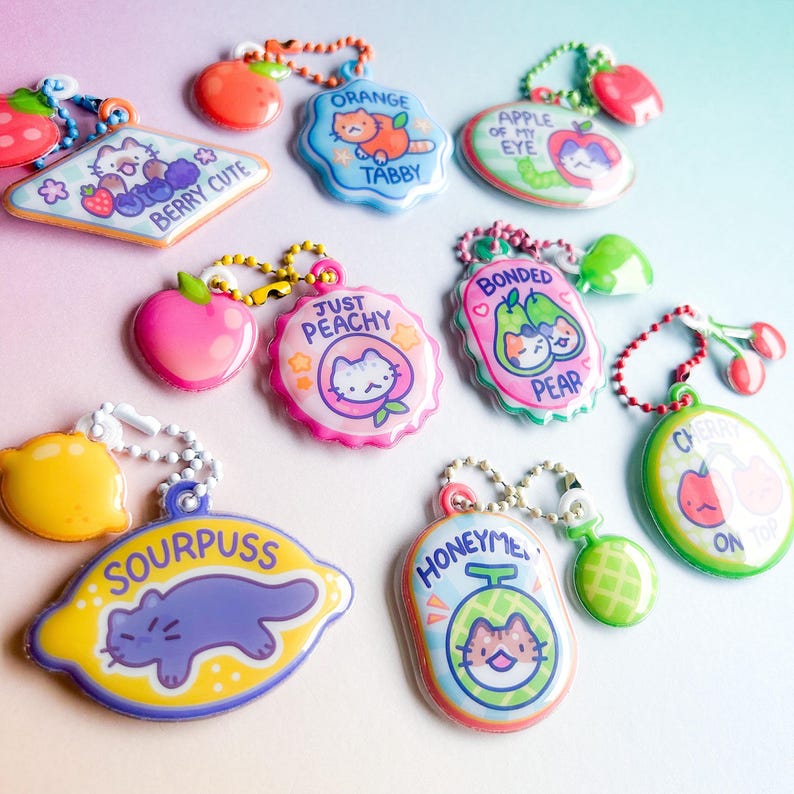 May include: A collection of colorful cat-themed keychains with fruit designs. Keychains include phrases like "Berry Cute," "Orange Tabby," and "Just Peachy." Each keychain has a metal chain and a fruit-shaped charm, such as a strawberry, apple, or lemon.