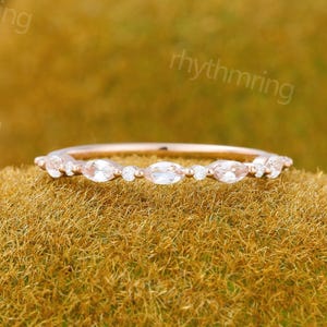 May include: A rose gold ring featuring a line of small, oval white gemstones. The ring sits on a textured, moss-green surface. The design is simple and elegant, suitable for everyday wear or special occasions. The ring is likely made of gold.