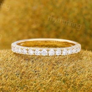 May include: A gold band ring featuring a continuous row of small, clear gemstones. The ring is set against a textured, golden-brown background, creating a contrast that highlights the ring's sparkle and design. The ring is a delicate piece of jewellery.