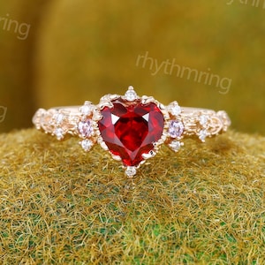 May include: A gold-toned ring featuring a heart-shaped, deep red gemstone as the centerpiece. The ring is adorned with smaller, clear and purple gemstones, and intricate detailing along the band. The ring is set on a textured, green surface.