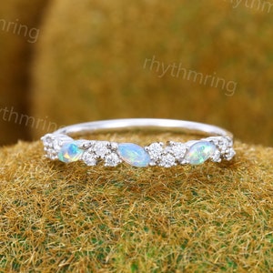 May include: A silver ring with a band of opals and small diamonds. The opals are oval-shaped and display a blue and green shimmer. The diamonds are set between the opals. The ring is shown on a textured, moss-like surface.
