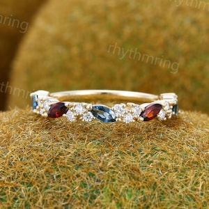 Pode incluir: Um delicado anel de banda de ouro com uma fila de topázios azuis em forma de marquesa, granadas vermelhas e diamantes brancos redondos alternados.