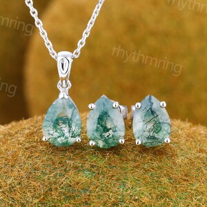 May include: A silver necklace with a teardrop-shaped pendant and matching stud earrings. The pendant and earrings feature a green and white moss agate stone. The jewelry set is displayed on a textured, brown surface.